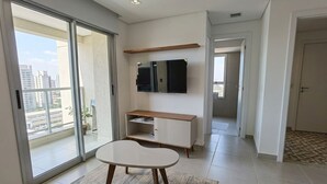 A modern living room with a sliding glass door to a balcony, a flat-screen TV mounted on the wall, and a minimalist furniture set.