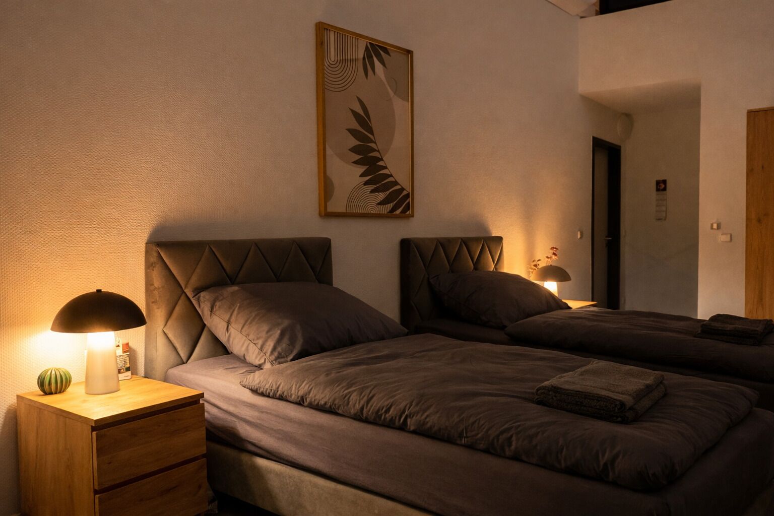 Une chambre avec deux lits, une table de chevet avec une lampe, une œuvre d’art encadrée au mur et une porte en bois.