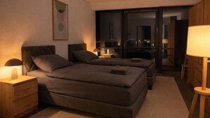 Un dormitorio moderno con dos camas, una mesita de noche, una lámpara y vista a la ciudad de noche.
