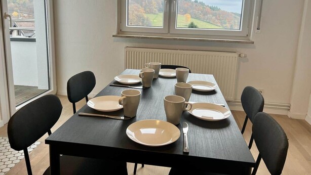 A dining table set with plates and cups, black chairs, and a view of a forest through a window.