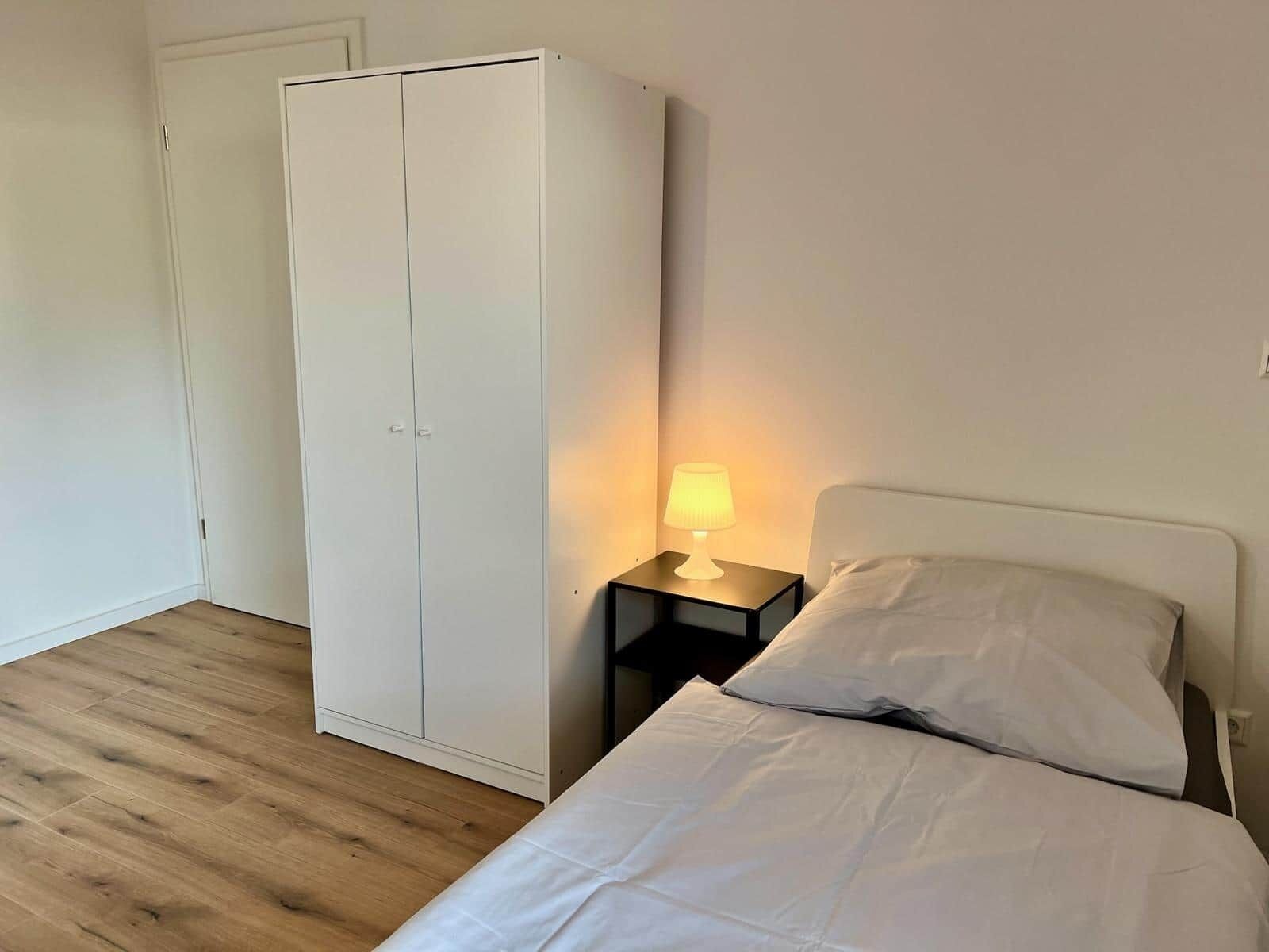 A bedroom with a bed, a nightstand with a lamp, and a wardrobe.