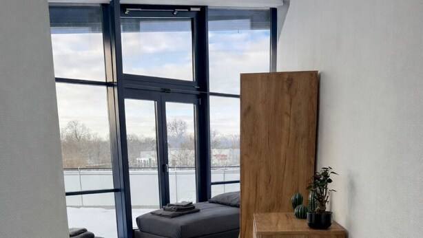 Ein modernes Interieur mit einem großen Fenster, einer Holzkommode, einem dunklen Sofa und einem Sessel.