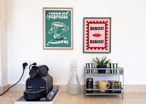 Eine Küche mit einer Kaffeemaschine, einem gerahmten Poster von einer Kaffeetasse und einem Regal mit einer Pflanze und Tassen.