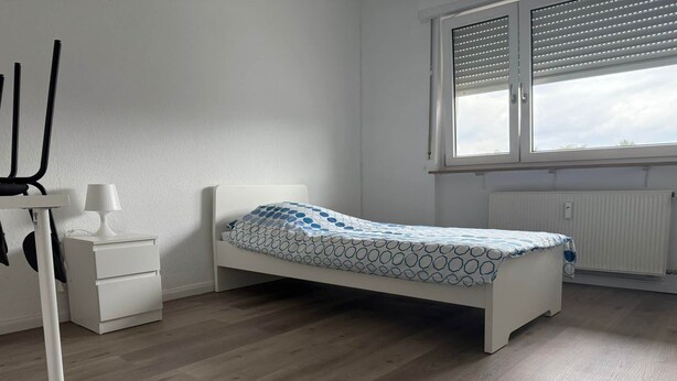 Ett enkelrum med en vit säng, ett nattduksbord med en lampa, ett element och ett fönster med persienner.