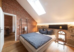 A bedroom with a lofted ceiling, exposed brick walls, a bed with a grey blanket, a small table, and a lamp. - Elegant Retreat: Private Sauna, Whirlpool Bath and Fireplace - Last Minute (Grossharthau)