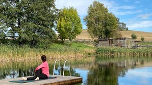 Eine Person sitzt auf einer hölzernen Anlegestelle, die sich in ruhiges Wasser erstreckt, umgeben von Bäumen und einer Grünfläche.