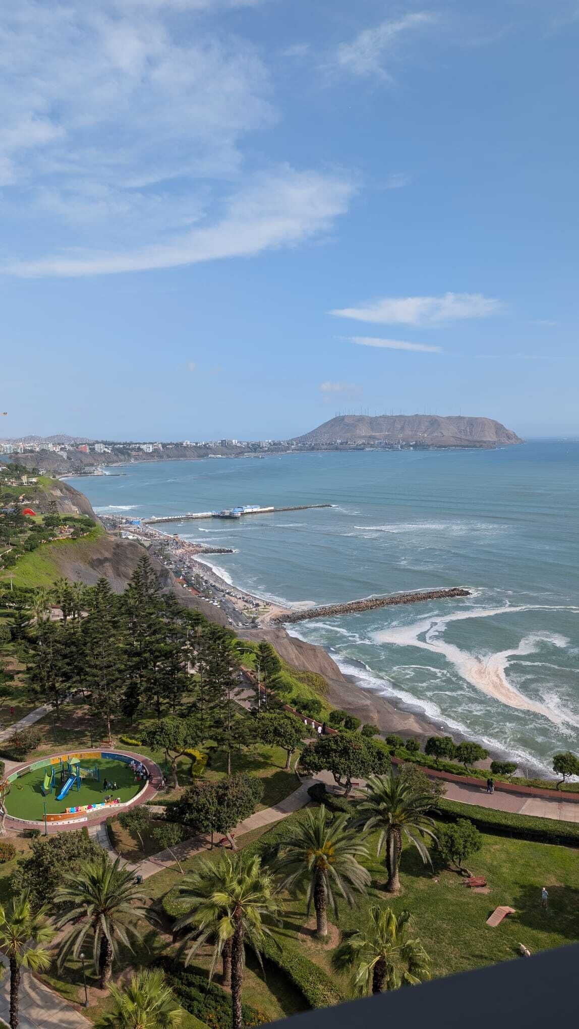 A coastal landscape with a beach, a rocky outcrop, and a park with palm trees.