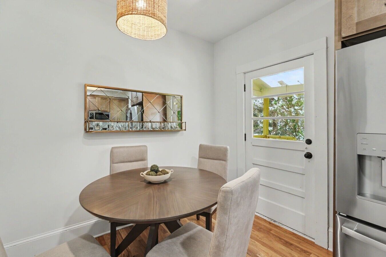 A dining area with a round table, chairs, a bowl of fruit, a mirror, and a door leading to a balcony.