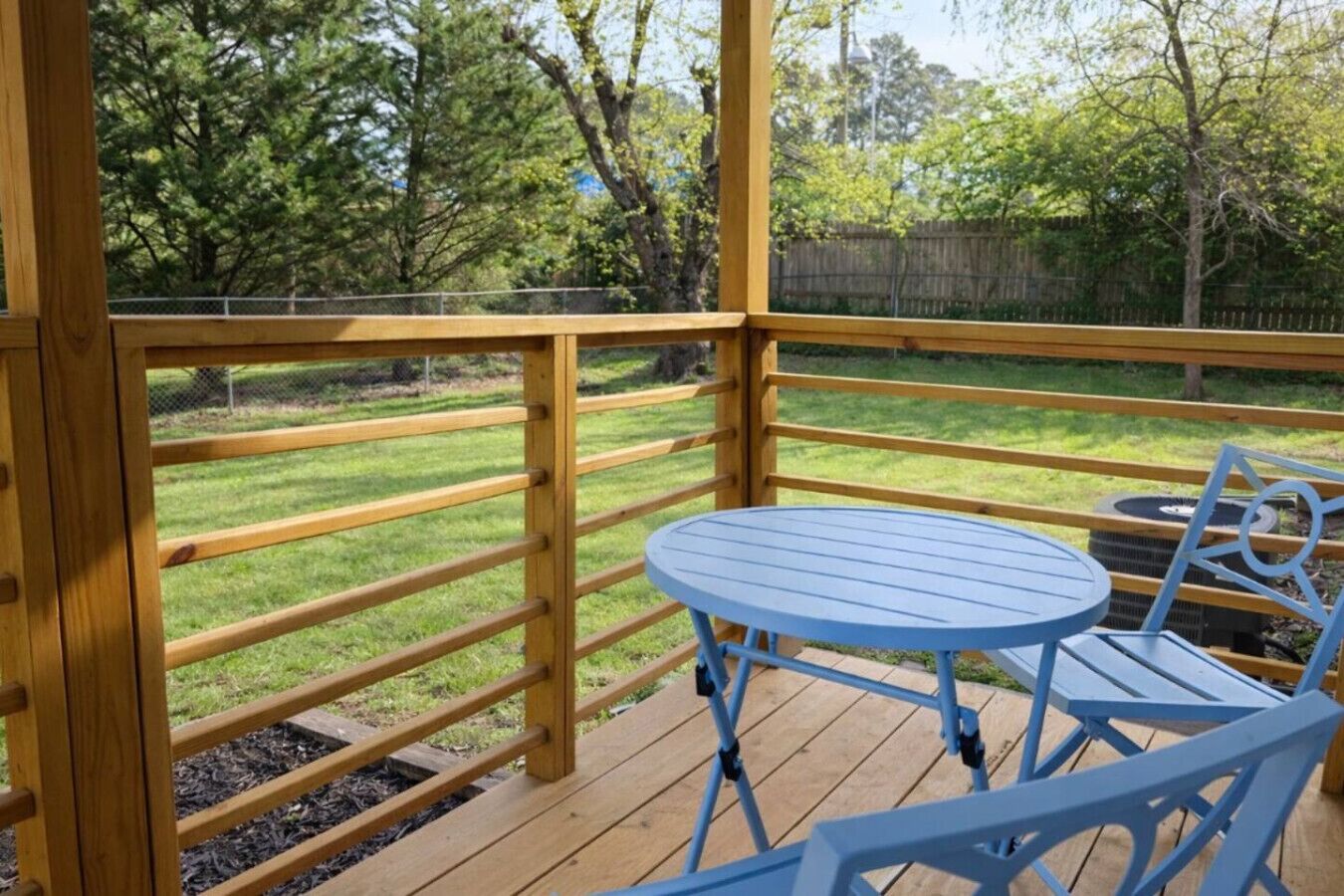 A wooden deck with a blue table and chairs, surrounded by a wooden railing and a grassy yard.