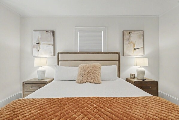 A bedroom with a large bed, two bedside tables with lamps, a window with blinds, and two wall-mounted paintings.