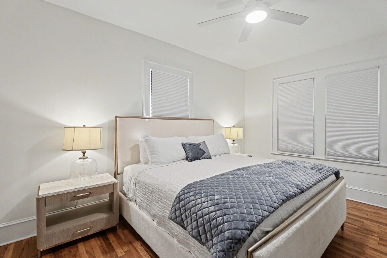 Un dormitorio con una cama, mesitas de noche, lámparas, ventilador de techo y persianas.
