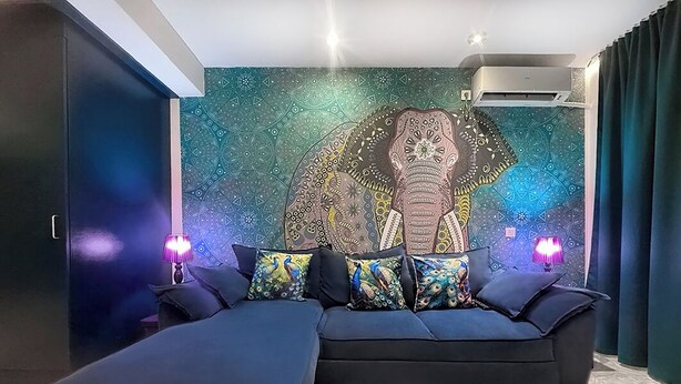 A modern living room with a large sofa and ottoman, a peacock-themed wall tapestry, and a ceiling-mounted air conditioning unit. - BLUE PEARL 2-BEDROOM LUXURY MAISONETTE IN PANORAMIC ROOF COMPLEX (Balchik)