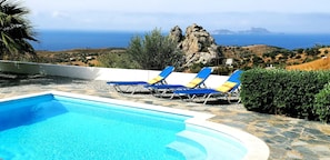 Ein Pool mit klarem, blauem Wasser, umgeben von Steinplatten und zwei Liegestühlen mit blauen und gelben Kissen.