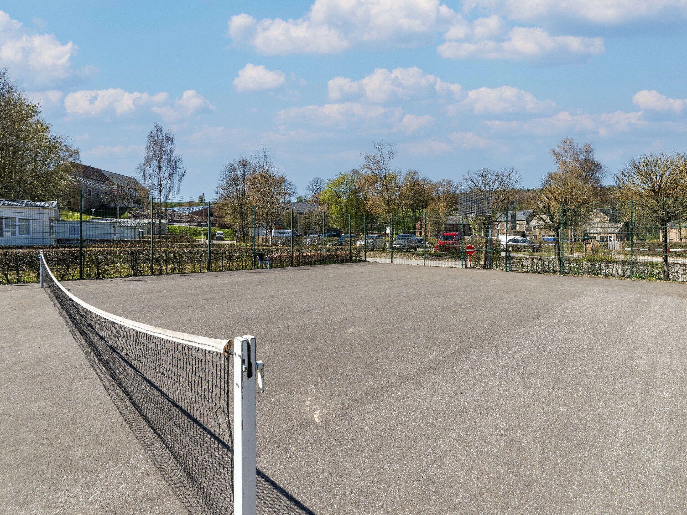 Una cancha de tenis al aire libre con una red, rodeada por una cerca, y casas al fondo.