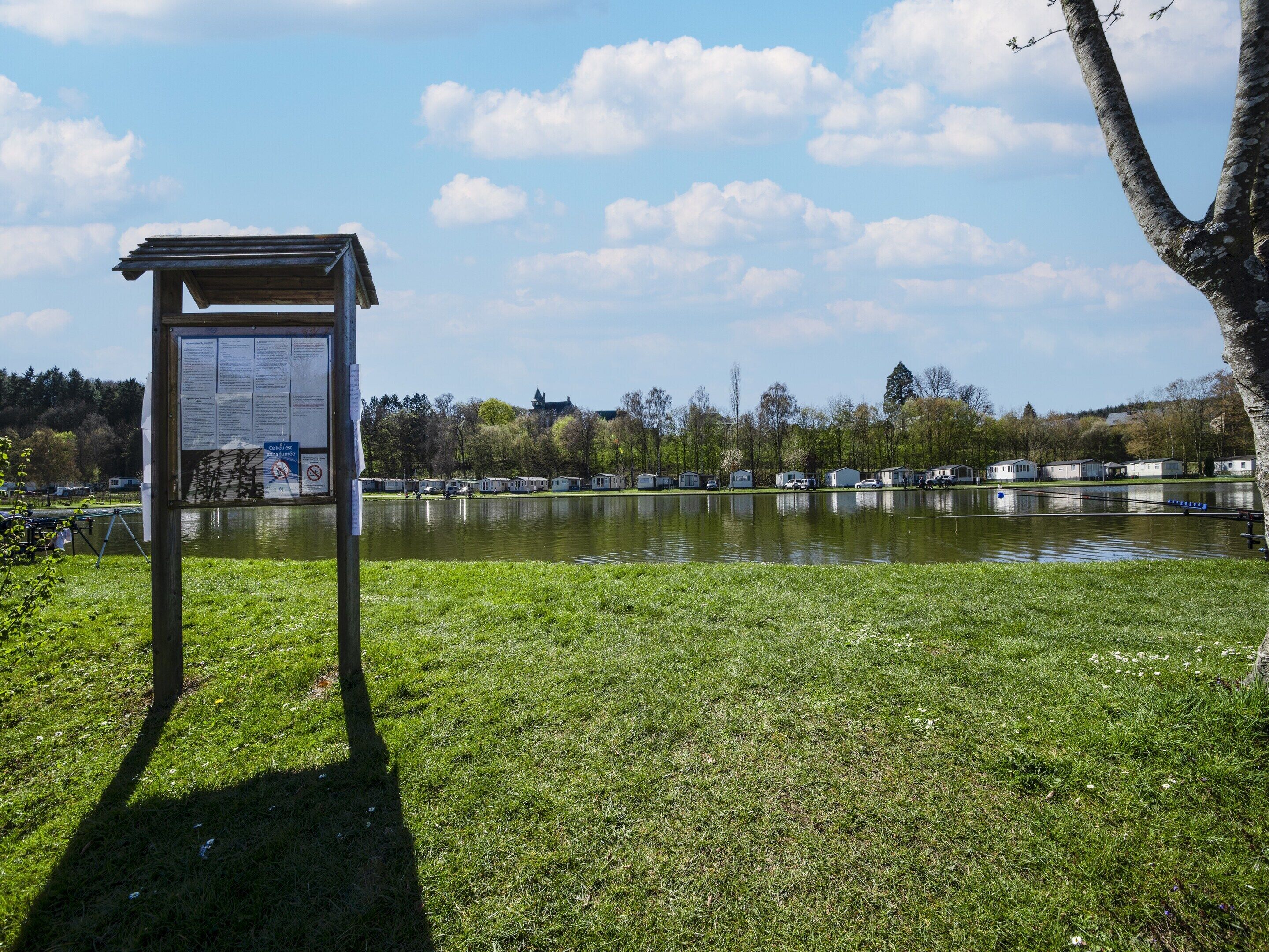 A lakeside view with a wooden information board, grassy areas, and row of mobile homes in the background.