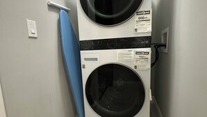 Ein Waschraum mit zwei nebeneinander gestapelten Frontlader-Waschmaschinen und Trocknern, einem blauen Bügelbrett und einem Wäscheeimer mit Waschmittel.