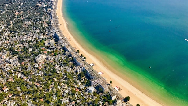 Luchtbeeld van een kustplaats met een lang strand, een dichtbevolkt woongebied en een heldere blauwe zee.