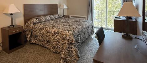 Una camera d'albergo con un letto, una scrivania, una sedia, un comodino, due lampade e una finestra vista alberi.