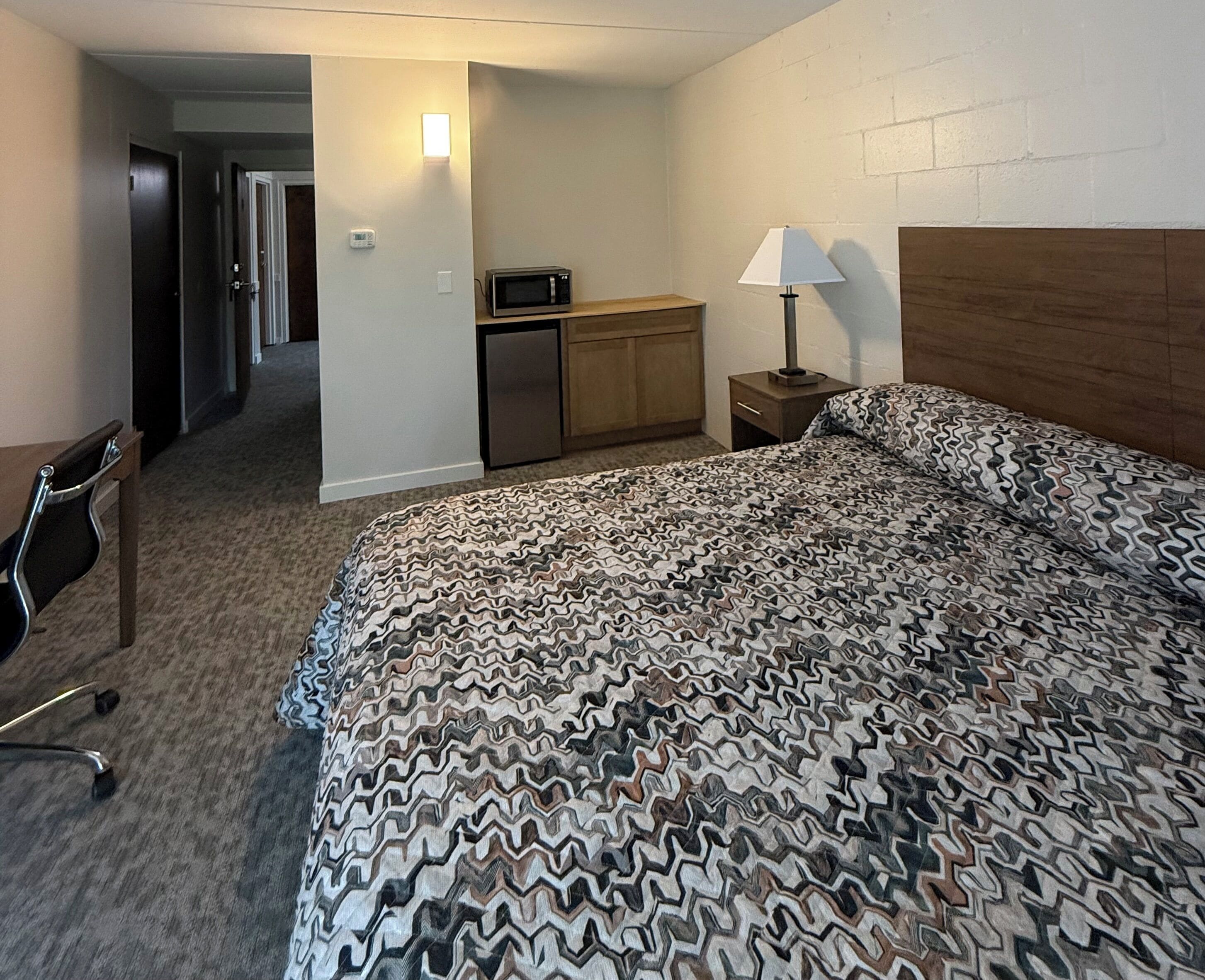 A hotel room with a bed, a desk, a chair, a microwave, a lamp, and a wooden headboard.