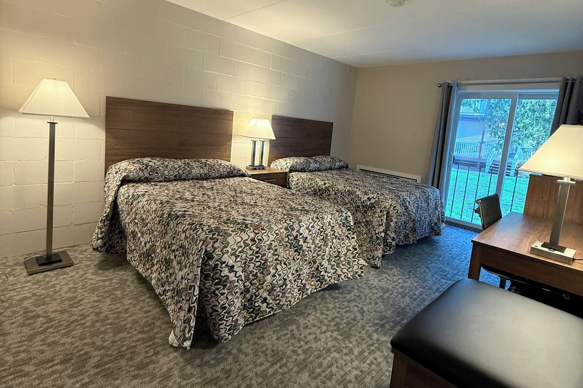A hotel room with two beds, a desk, two lamps, and a view of the outdoors.