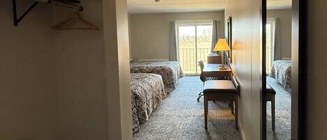 Habitación de hotel con cama, escritorio, silla, lámpara, espejo y perchero.