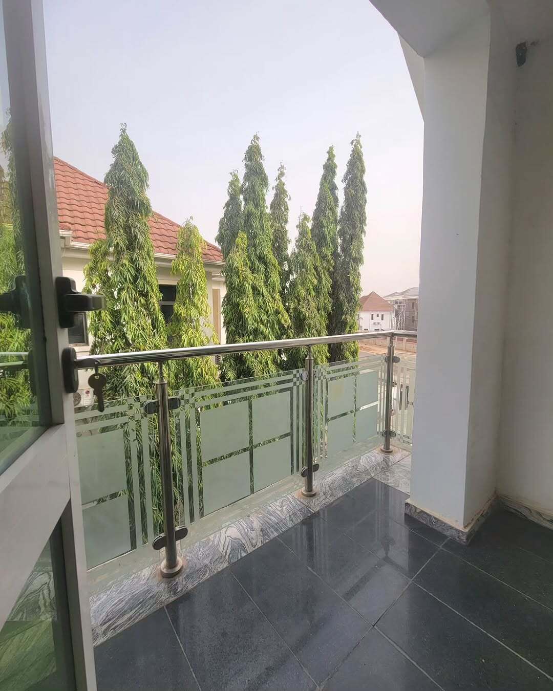 A balcony with a glass railing, a view of trees, and a tiled floor.