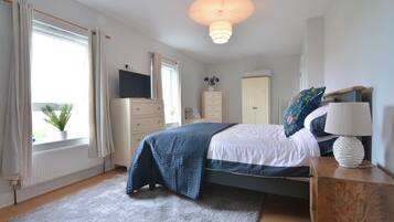 A bedroom with a bed, bedside table, lamp, dresser, TV, and window with curtains.