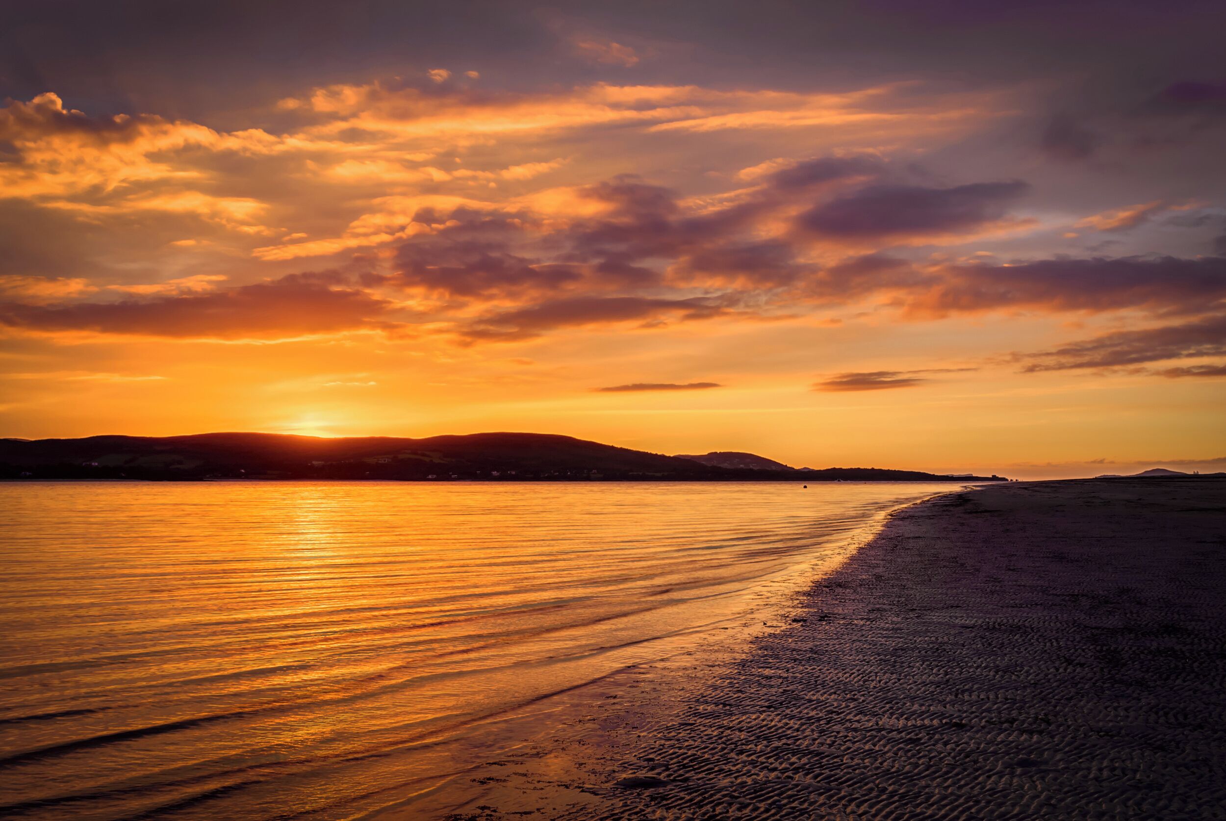Uma praia com um pôr do sol, águas calmas e colinas ao longe.