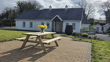 A house with a paved patio, a wooden picnic table with a vase of flowers, and a well-maintained lawn.