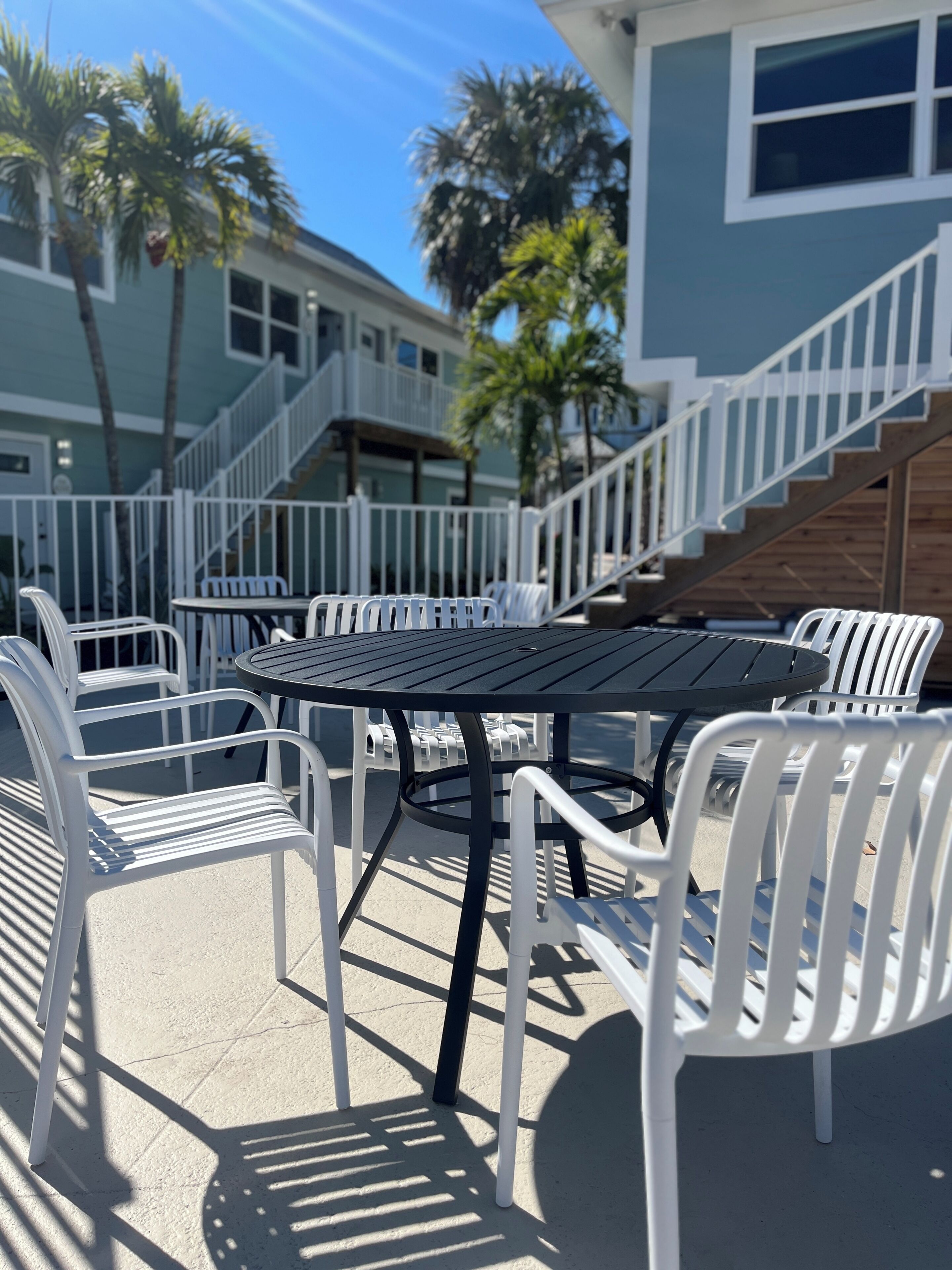 An outdoor patio with white plastic chairs and a table, a staircase, and a building with balconies.