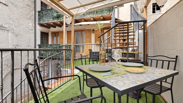 Un área de comedor al aire libre, techada, con una mesa preparada para dos personas, rodeada de sillas y una escalera que conduce a un nivel superior.