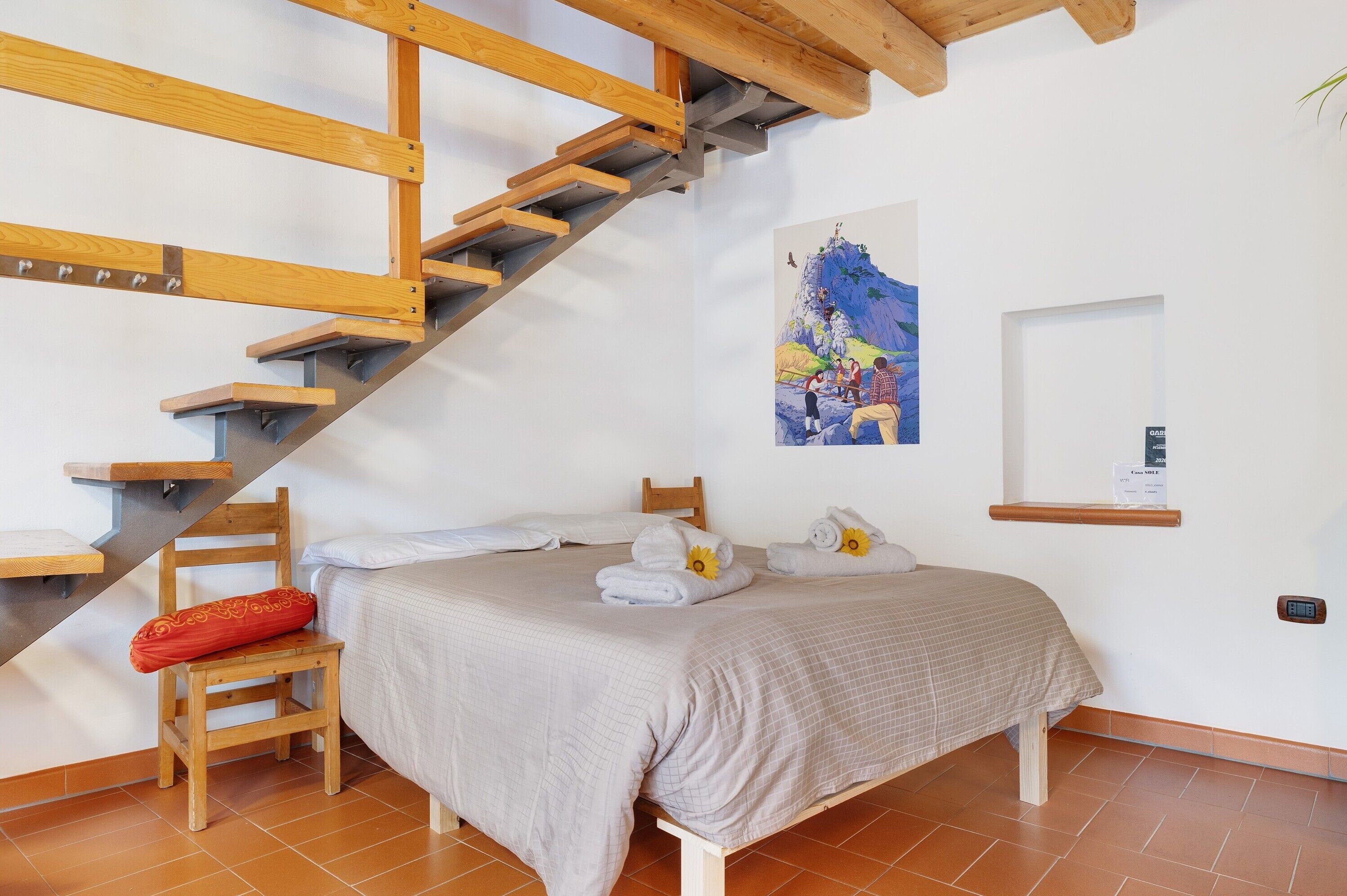 Ein Schlafzimmer mit einer Holztreppe, einem Bett mit einer beige farbenen Tagesdecke, einem Sessel mit einem roten Kissen und einem Gemälde an der Wand.