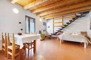 Una habitación con escalera de madera, cama con ropa blanca, una mesa de comedor con sillas y una pequeña zona de cocina.
