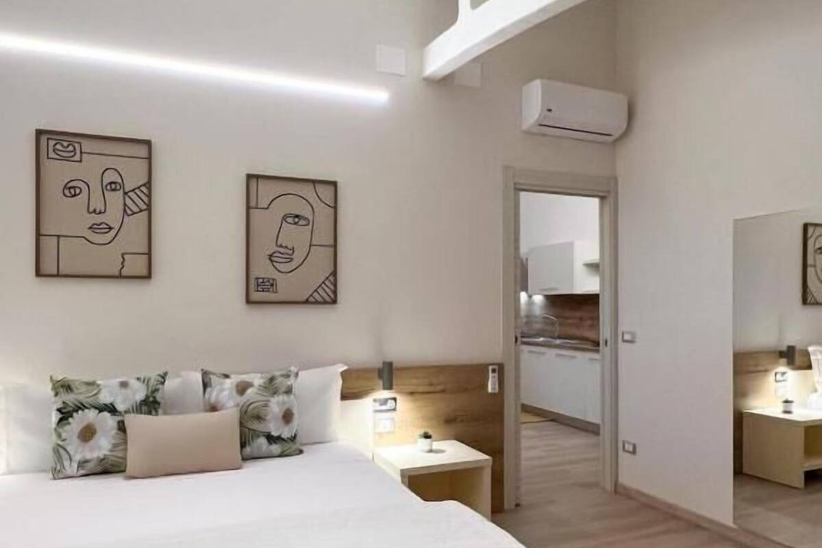A modern hotel room with a bed, a nightstand, and a mirror. There are two wall art pieces and an air conditioning unit.