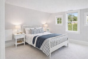 A bedroom with a white bed, two bedside tables with lamps, a radiator, and a window with a view of greenery. - Falmouth Foreside Cottage Escape - Luxe Kitchen + Walk to the Water! (Falmouth)