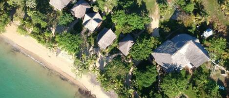 Vista aérea de un resort frente a la playa con cabañas de techo de paja, palmeras y una playa de arena.