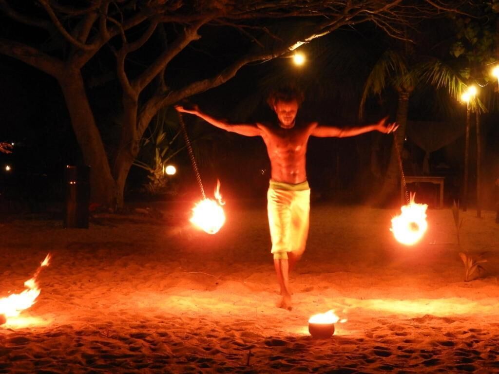 Une personne effectue un numéro avec du feu sur une surface sablonneuse, entourée d’arbres et éclairée par une lumière discrète.
