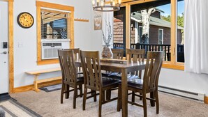 A dining area with a wooden table and chairs, a chandelier, a clock, and a window with a view of a cabin.