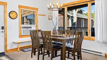 A dining area with a wooden table and chairs, a chandelier, a clock, and a window with a view of a cabin.