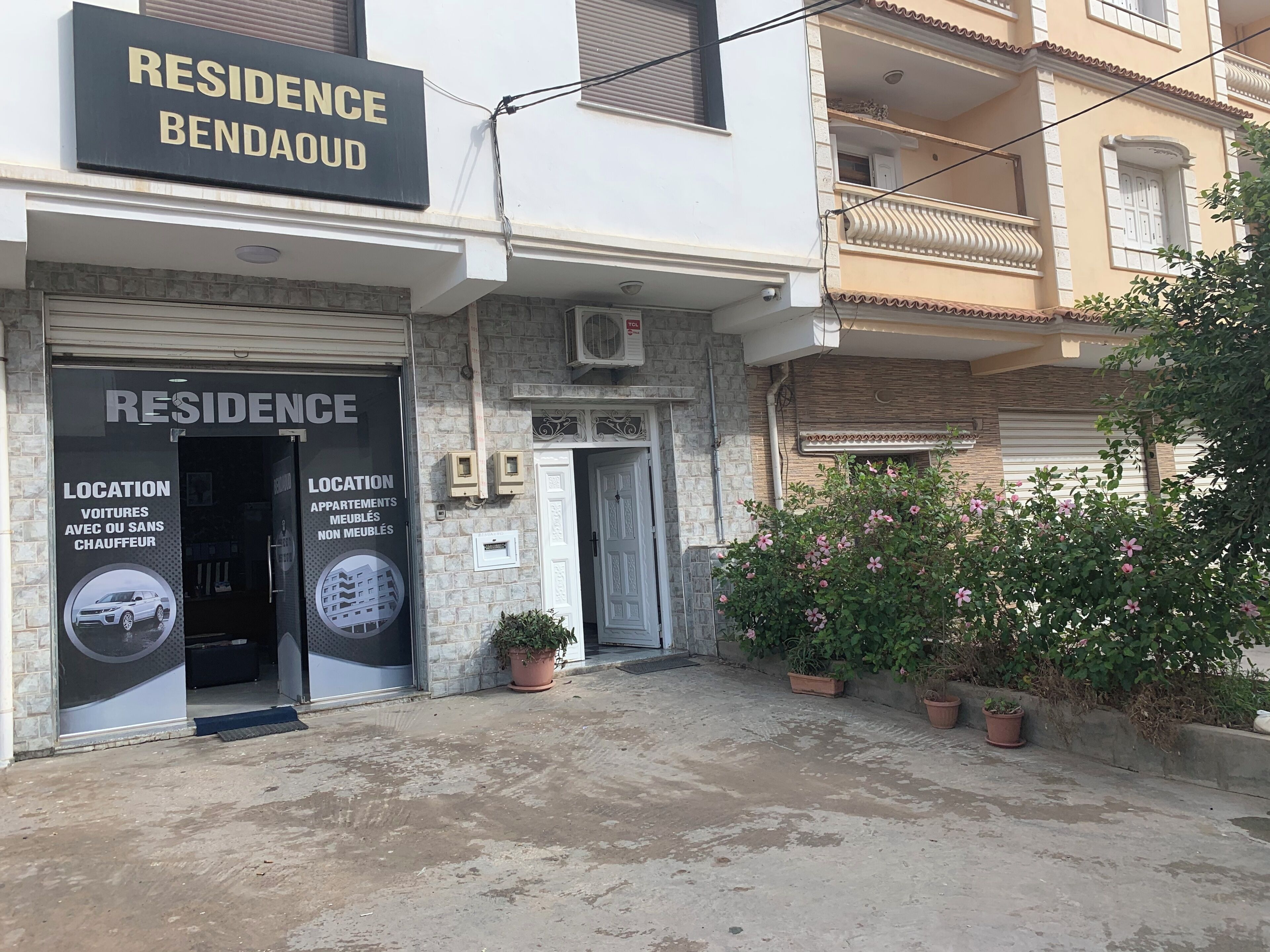 A building with a sign reading 'RESIDENCE BENDAOUD' and a door labeled 'RESIDENCE.' There are potted plants and a small garden area in front.
