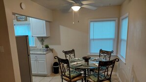 Comedor con una mesa redonda, cuatro sillas, un ventilador de techo y una cocina al fondo.