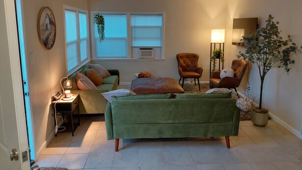 Un dormitorio con un sofá verde, dos sillones, una mesita con una lámpara, una planta en maceta y un cuadro en la pared.