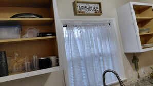 Una cocina con estantes de madera, una ventana con cortina, un grifo y un cartel que dice 'FARMHOUSE'.