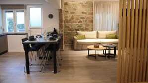 A dining area with a table and chairs, a sofa with cushions, a small round table, and a stone wall. - CozyFlat 88m2 very spacious for long stays, fully equipped, with private parking (Mitilini)
