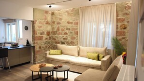 A living room with a stone wall, a sofa, two small tables, and a kitchen area with a counter and cabinets. - CozyFlat 88m2 very spacious for long stays, fully equipped, with private parking (Mitilini)