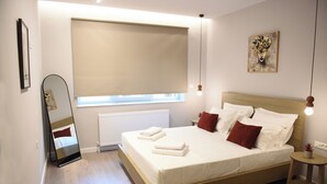 A bedroom with a bed, a window with a blind, a bedside table with a lamp, and a wall-mounted artwork. - CozyFlat 88m2 very spacious for long stays, fully equipped, with private parking (Mitilini)