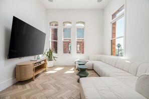 Una sala de estar moderna con un televisor de pantalla plana grande, piso de madera con diseño espiga, un sofá blanco y un ventanal con vista a un edificio de ladrillo.