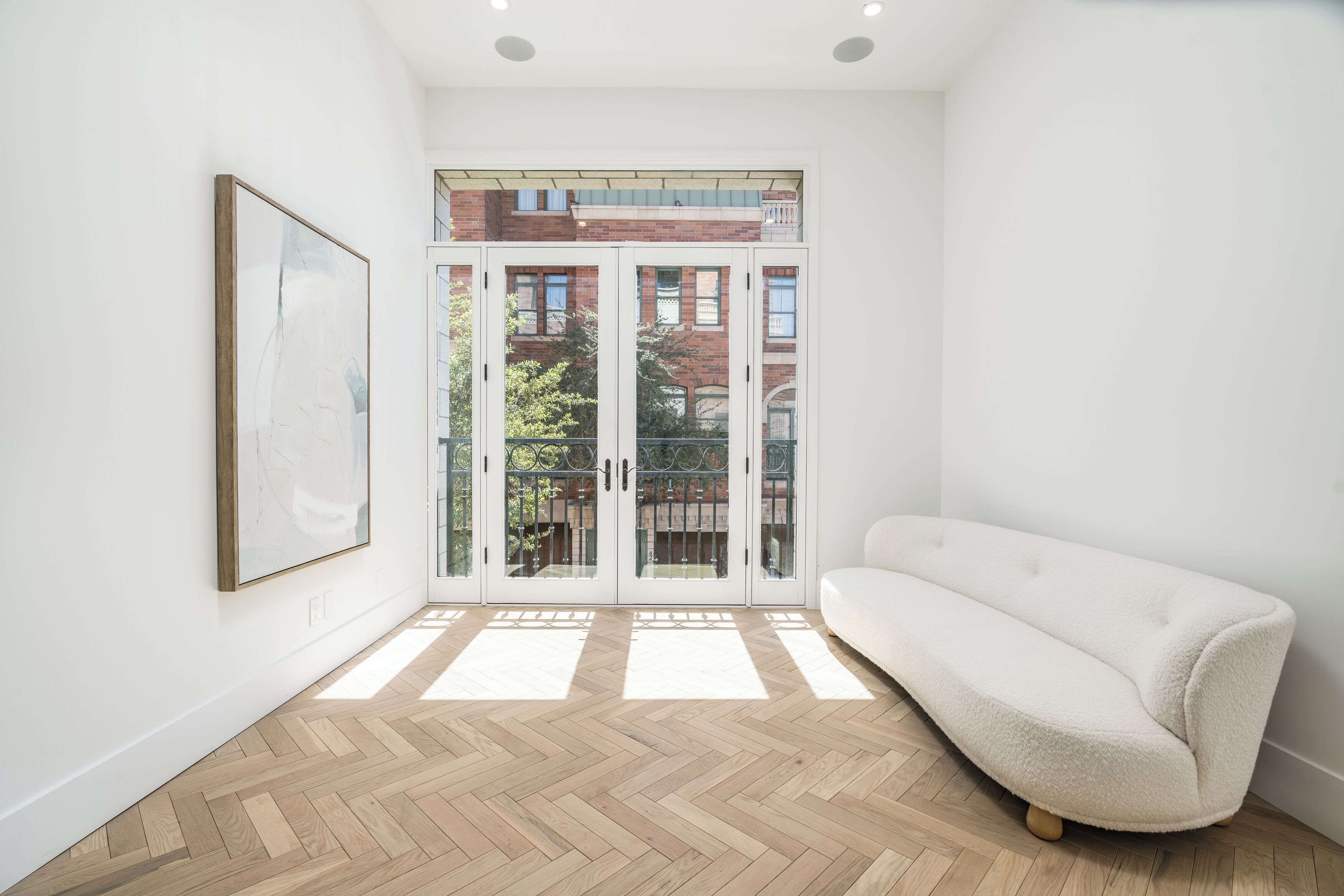 A room with a herringbone floor, a white sofa, a framed picture on the wall, and a glass door leading outside.