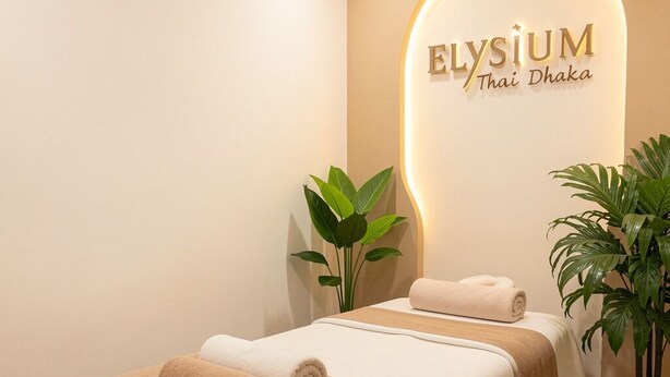Une salle de spa équipée d’un lit, de serviettes, d’une plante en pot et d’un mur sur lequel est inscrit le nom « Elysium Thai Dhaka ».