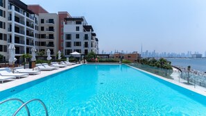 En pool med solstolar och parasoller, en stadsbild i bakgrunden och en klar himmel.